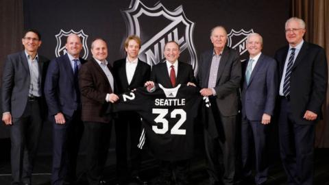 NHL exeuctives hold up a black jersey with Seattle 32 written on the back in white