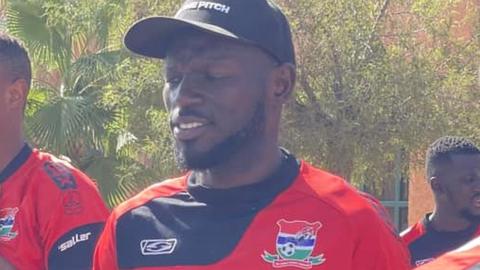 Gambia captain Omar Colley walks along wearing a team training shirt and baseball cap