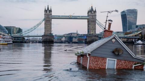 A replica of a British house floats in front of Tower Bridge on the river Thames