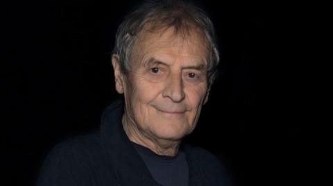 A man with grey hair wearing a navy blue top and jacket, against a black background. 