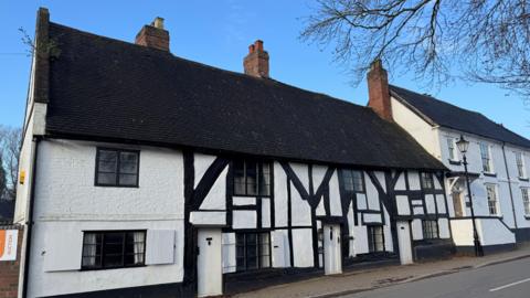 A timber-framed, black and white building. There is another, taller, white building next to it and they are both set against the backdrop of a blue sky.