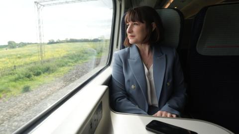 Rachel Reeves looking out of a train window. She is seated and looking to her right. She has dark hair and is wearing a pale blue jacket and white blouse. A mobile phone is placed on the table in front of her. 