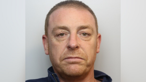 A police custody photo of Darren Jones. He has very short ginger hair and stubble. He is wearing a dark blue zip-up jumper.