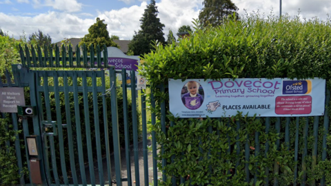 A banner which reads 'Dovecot Primary School - Learning together, growing together - places available'. It is hung on a green fence outside the school and is surrounded by a large green bush.