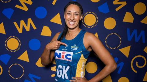 Geva Mentor smiling at the camera in a blue netball kit and pointing to the Leeds Rhinos badge on her chest 