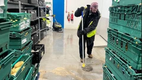 A man wearing a hair net and high viz vest mops up flood-water from a warehouse floor. Green crates of food are stacked up either side of him. Behind him, another person is visible using a flood vacuum