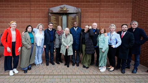 Large group picture of about a dozen Hillsborough campaigners including Margaret Aspinall with David Lammy, Steve Rotheram and Andy Burnham. They are lined up outside the memorial listing the names of 97 victims on the wall of Anfield stadium.