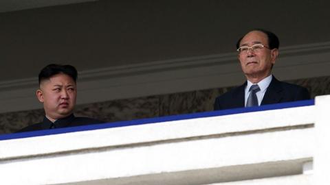 Kim Jong Un standing on a balcony glancing at his side towards Kim Yong Nam