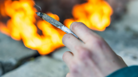 A hand is holding a rolled up cigarette. A fire outside is out of focus. 