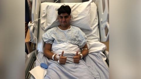 A young man with dark brown hair is in a blue and white hospital gown sitting on a hospital bed. He has his thumbs up and is smiling at the camera