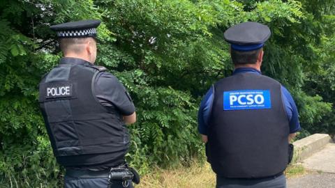 Two officers in uniforms and black hats stood facing forwards. On the backs of their uniforms one reads 'POLICE' the other reads 'PCSO' stood in front of police taper by a bridge in a leafy area.
