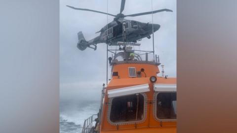 A grey helicopter hovers over an orange lifeboat in the middle of the sea during a rescue operation. It is a cloudy day. A man wearing a yellow helmet is on the helm of the boat.