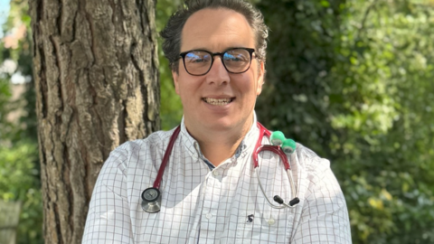 The image shows Dr Ed Klaber standing outdoors in front of a tree with greenery in the background. Dr Klaber is wearing a white and black checked shirt. He is wearing glasses and has a stethoscope around his neck.