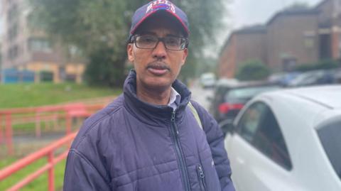 A man with a thick winter coat, baseball cap and glasses stands on the road next to cars parked up the pavement.