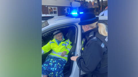 An elderly woman sits in a police car with the door open, she wears a hi-vis police jacket and police hat and is smiling at a female police officer who is helping her out of the car 