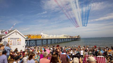 Crowds of people sat on Paignton beach admiring a Red Arrows display overhead, the blue sea in the distance alongside Paignton pier, on a sunny day.