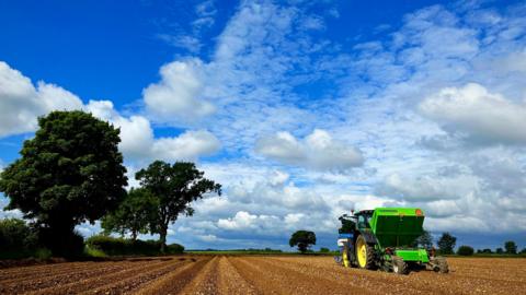 THURSDAY - A green tractor at Woodstock drives through an earth field with a blue sky and green trees in the hedgerow