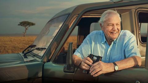 Sir David Attenborough leans onto an open car door whilst holding a pair of binoculars.
