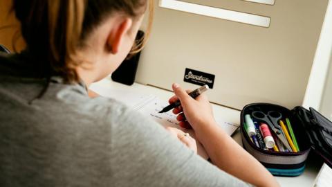 A stock image of a young girl pictured from the back, sitting at a desk doing homework. She is holding a black pen and writing in a notebook. She has a pencil case next to her, with pencils, pens and scissors.