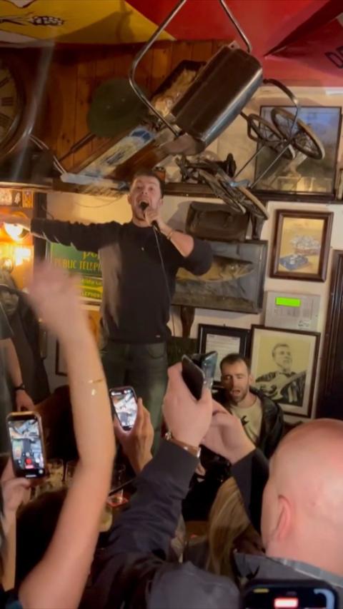 Eddie Keogh stands on a pub table wearing a grey shirt and green trousters with his hand extended holding a microphone. There are several people below him taking videos on their phones.