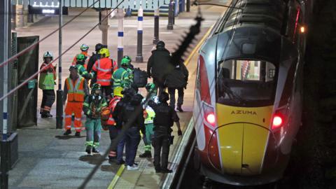 A group of people in high-vis green and orange vests and jackets crowd on a platform beside a train. It is after dark.