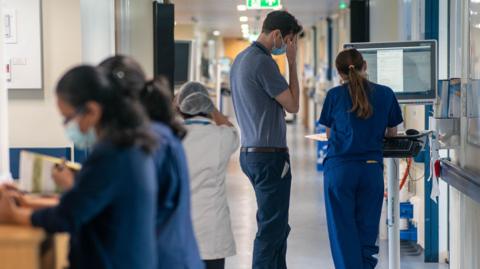 Nurses and a doctor looking at computers and read notes on a hospital ward. The doctor has his hand over his face 