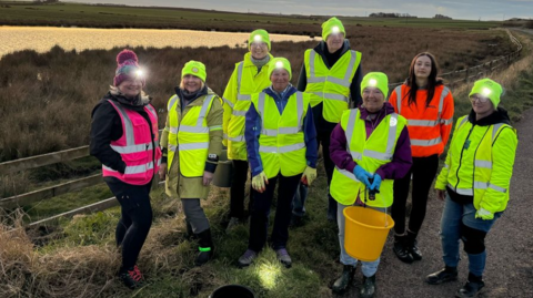 A group of people wearing hi-vis jackets and head torches are standing by the side of the road. One is carrying a bucket, behind them is a stretch of coastline.