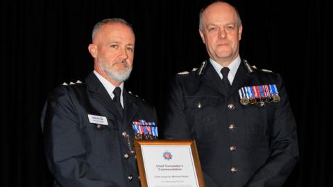 Two police officers in uniform are smiling while standing next to each other and holding a framed award certificate which says 'chief constable's commendation' and has the name 'chief inspector Michael Parker' written on it. The man on the left has short grey hair and a beard while the man on the right has receding grey hair. They both have a number of medals on their jacket.