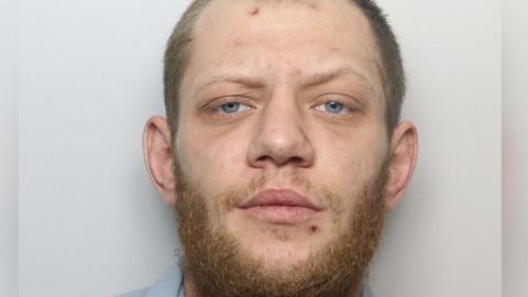 Ryan Surtees is facing the camera in a police photograph. He has shot brown hair and a short brown beard and is wearing a blue dressing gown