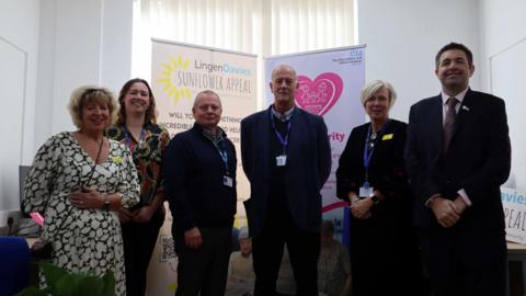 Three women and three men standing in a room. There are two large advertising board behind them. One reads "Lingen Davies sunflower appeal" and the other reads "SaTH charity". 