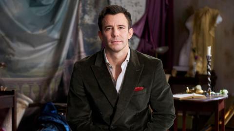 A man with short brown hair, actor Luke Thompson, is wearing a white shirt and dark grey tweed suit with a red handkerchief in the top pocket. Behind him is a fabric covering a wall with a desk and ornate candlestick and candle. 