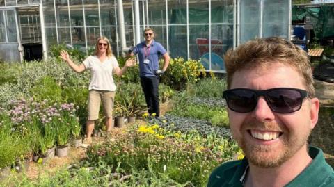 People stand and smile while standing in among lots of pots of plants sprouting various flowers close to a green house. 