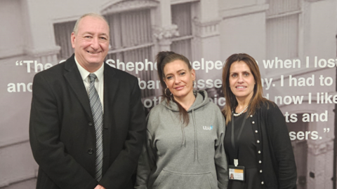 Councillor Steve Evans, Lindsey and Shaz stand in front of a wall poster for the Good Shepherd charity. Steve is wearing a dark suit, Lindsey is in a grey hoodie and Shaz has a dark top and cardigan on. The poster is in between two wooden doors.