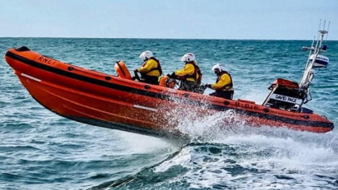 The image shows an RNLI lifeboat speeding across the water. The boat is bright orange and carries three crew members dressed in yellow jackets and white helmets. 
