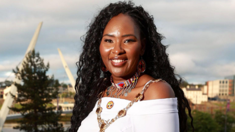 Shows Lilian Seenoi-Barr in a white dress wearing the mayoral chain and a colourful necklace and standing against the backdrop of the peace bridge.