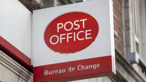 A red circle on a white sign. In the red circle are the words 'post office' in white. Behind the sign are brick walls.