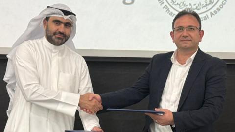Dr Mamoom Al Deyab is wearing a suit jacket with a white shirt, while Dr Ahmad Taqi is wearing a white ghutra and egal with white shirt. They are posing for the picture while shaking hands