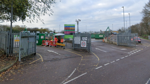 The entrance of the recycling and household waste site which has a silver fence at the perimeter. 