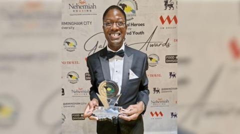 Karl Brown wearing a black suit with a black bow tie and glasses. He is at the award show, standing against a white banner with the logos of several universities and community groups. He is holding the metal award in both hands and smiling at the camera.