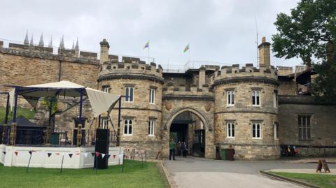 A picture of the gate house of the castle, with its turrets and castle walls. A temporary band stand structure is on the lawn in the left of the picture.