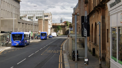 A view of a street. Bus shelters are on either side of the road, with a department store building on one side. Buses, a van and a blue car are visible on the road itself.