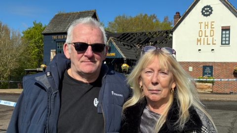 Mark and Carol Wenn standing in front of a police cordon at The Old Mill pub in Thorpe Marriott. Standing on the left is Mark who is wearing a black T-shirt and blue jacket. He is also wearing a pair of sunglasses over his eyes. Carol, on the right, is wearing a black and white coat, she has blonde hair and a pair of glasses rested on the top of her head.