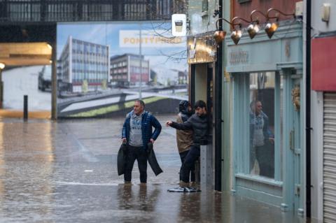 Flooding on Mill Street in Pontypridd. Three men are walking out of a shop into water that is ankle deep. In the background, a sign with Pontypridd is visible