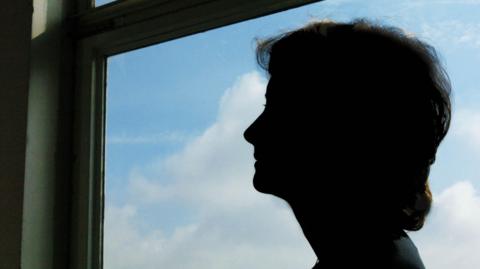 A silhouette of a woman looking out of a window, with a view of blue sky and white clouds.