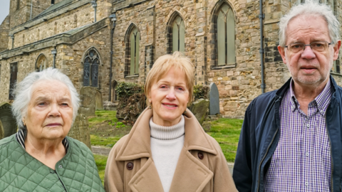 Parishioner Pat Baxter outside St Mary's with Churchwarden Christine Smithson and Bill Smithson. Pat Baxter has short white hair is wearing a green coat. Christine Smithson has short blonde hair, a light brown coat anf a white turtleneck. Bill Smithson is wearing a purple checkered shirt and a blue coat. He has short white hair and glasses.