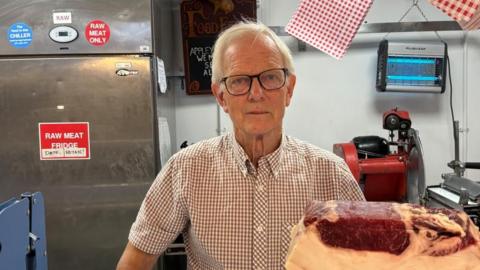 There is a man in a chequered button up shirt. He has black framed glasses on and short, thin grey hair. In front of him, to the right, is a piece of cured pork. Behind him to his right is a red meat cutter and an electric fly buzzer. To his back left, there is a big silver fridge with red stickers saying 'raw meat only' and 'raw meat fridge'