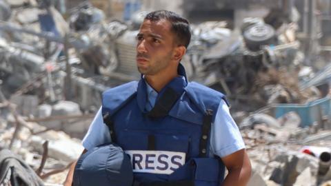 Al-Jazeera correspondent Anas al-Sharif stands among rubble. He is wearing a bulletproof vest with the words "press" on its front.