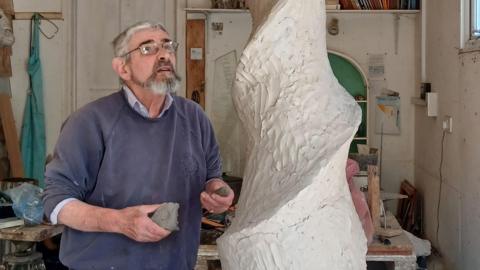 John working on the sculpture. He is looking up at a spiral made out of clay. He is wearing a blue jumper and glasses. He is in a studio.