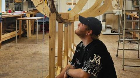 Jacob Beak, wearing a black t-shirt and cap, is looking up at a wooden boat-like structure. It is in a large workshop.