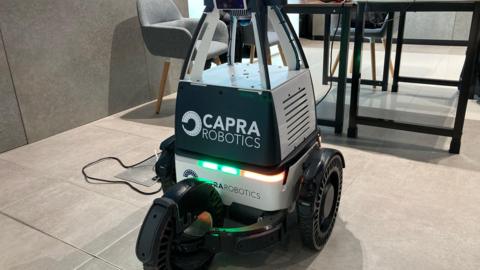 A robotic device is pictured on a tiled floor. It has three wheels and is black and silver. Printed on the front are the words "Capra Robotics"